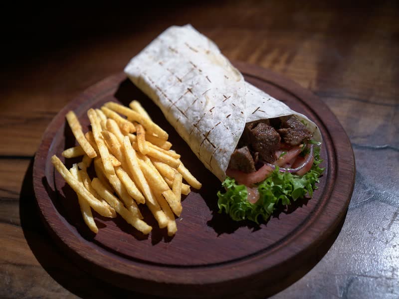 Een gegrilde durum-wrap met vlees, sla en tomaat, naast een portie friet op een houten plank.